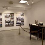 W nowoczesnej sali z białymi ścianami stoi stare biurko z eksponatami, na ścianach wiszą fotografie